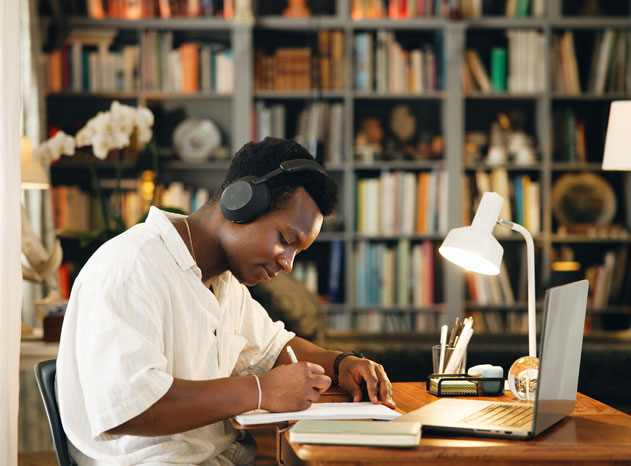 Hombre con auriculares escribiendo en una libreta junto a un portátil, frente a una estantería con libros.